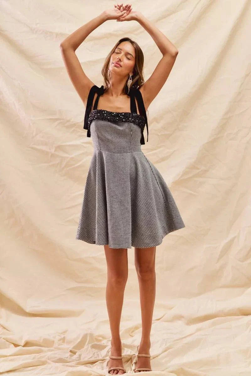 Woman wearing stylish houndstooth fit flare mini dress with black adjustable shoulder ties against beige backdrop