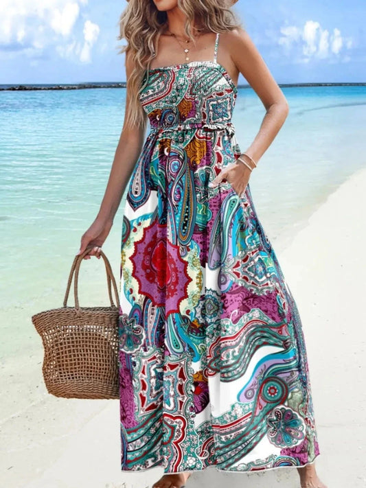 Woman wearing a colorful printed maxi cami dress with smocked waist detail on a beach, holding a woven handbag