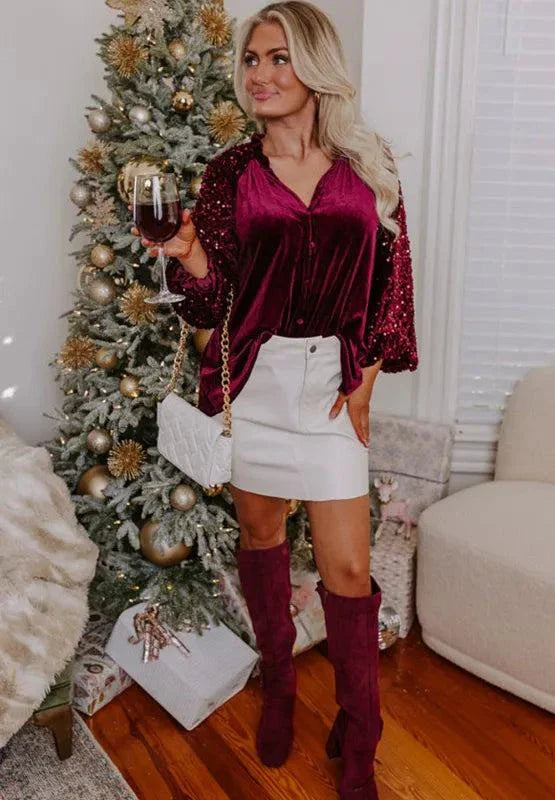 Woman wearing burgundy sequin notched long sleeve blouse with white skirt and matching boots near Christmas tree