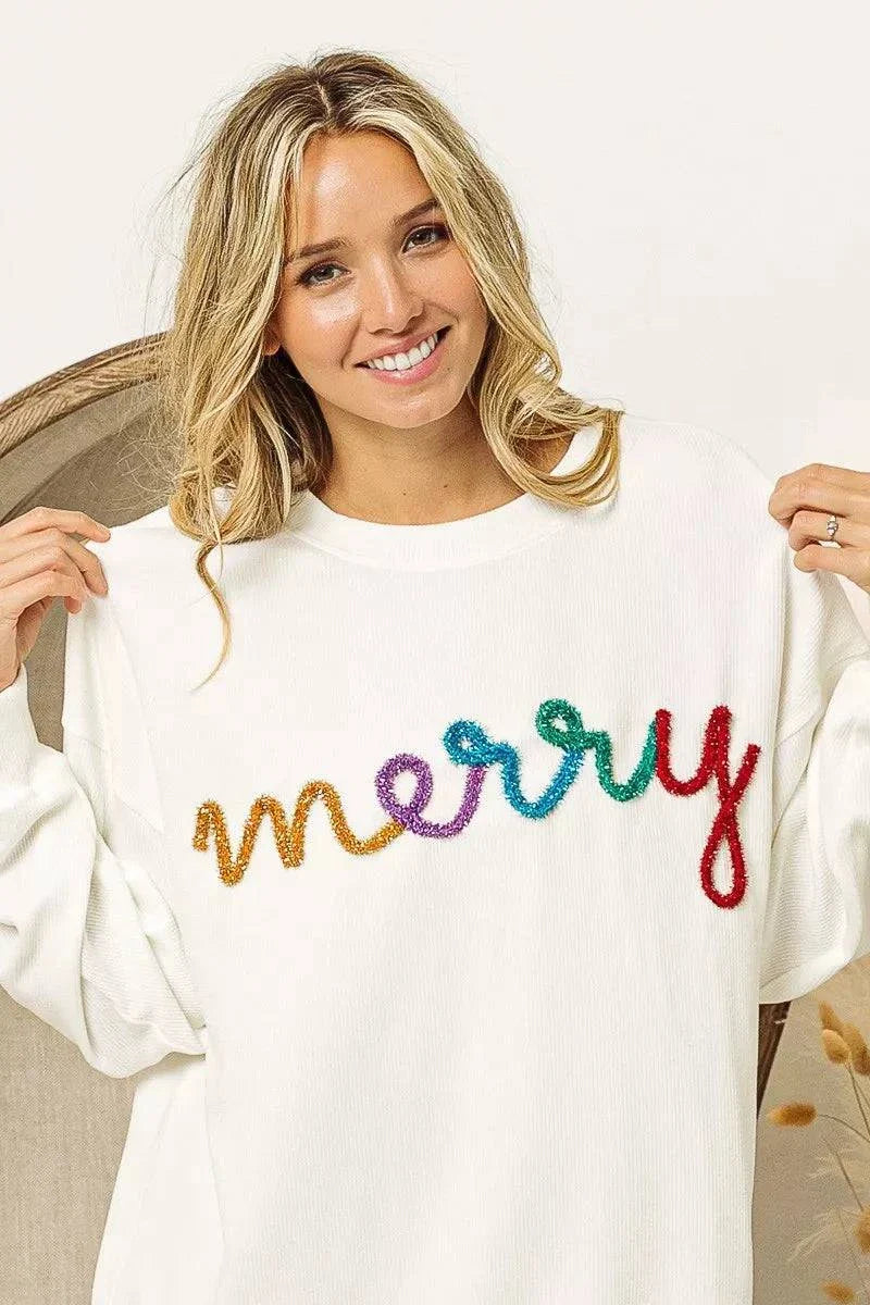 Woman wearing white drop shoulder sweatshirt with colorful Merry Tinsel Lettering across front